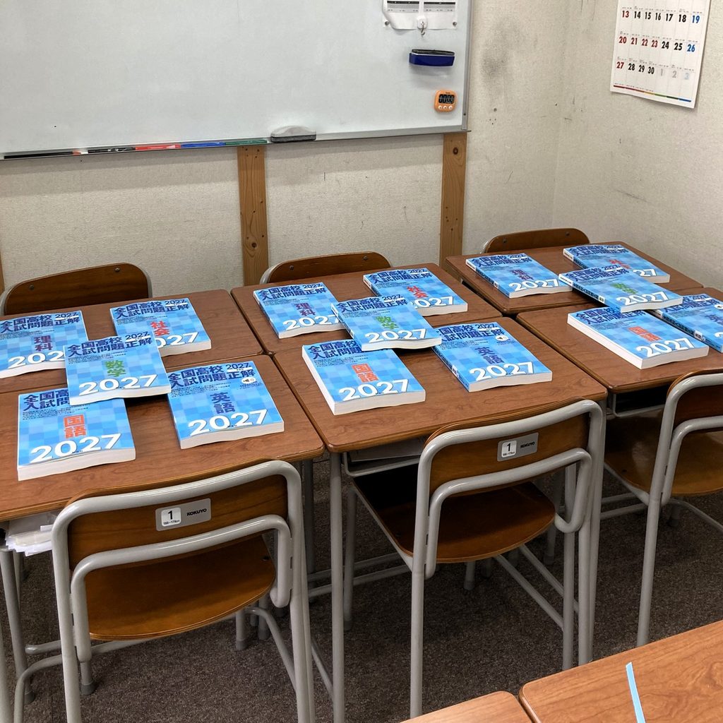 千尋進学塾の教室で、机の上に並べられた旺文社『全国高校入試問題正解 2027』。国語・数学・英語・理科・社会など各教科の青い問題集が複数の机に置かれている。中学3年生の四日市高校上位合格クラスでは、これらを使って全国の公立高校入試問題に取り組み、本格的な入試演習を進めている。